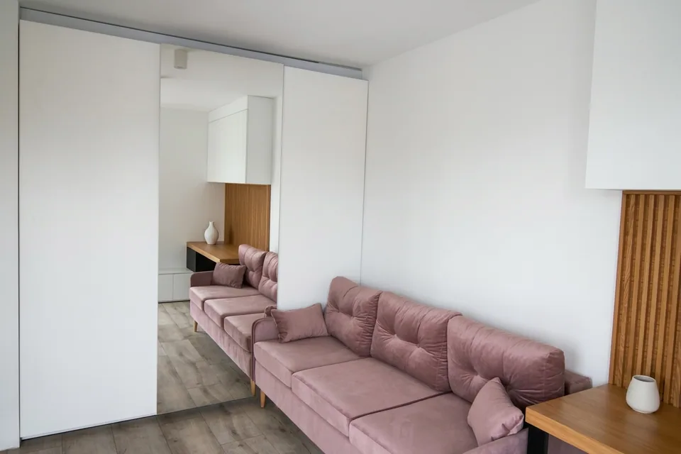 Różowa sofa pod jasną ścianą z wiszącymi szafkami